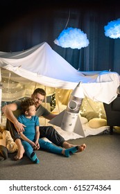 Father And Son Sitting Together In Blanket Fort And Playing With Toy Rocket