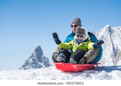 Father And Son Sledding During Winter Holiday. Happy Dad And Little Boy Playing With Snow Sled. Man With Smiling Child Sitting On Bobsledge In The Snow With Copy Space.