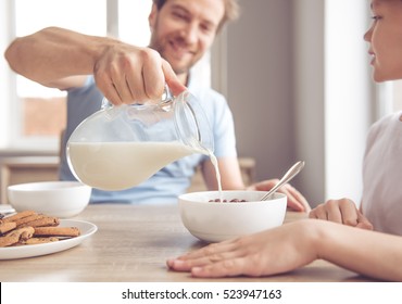 Father And Son Are Smiling While Having A Breakfast In Kitchen. Dad Is Pouring Milk Into Bowls