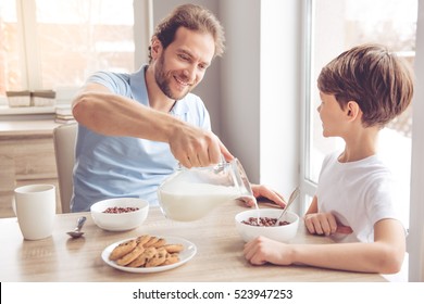 Father And Son Are Smiling While Having A Breakfast In Kitchen. Dad Is Pouring Milk Into Bowls