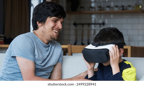 A father and son spending quality time at home. Boy wearing VR headset, experiencing immersive technology, while father watches and smiles. Modern family interaction and bonding. - Powered by Shutterstock