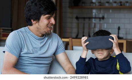 A father and son spending quality time at home. Boy wearing VR headset, experiencing immersive technology, while father watches and smiles. Modern family interaction and bonding. - Powered by Shutterstock