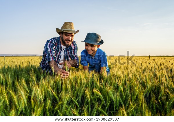 Father and son\
are standing in their growing wheat field. Father is teaching his\
successor about\
agriculture.