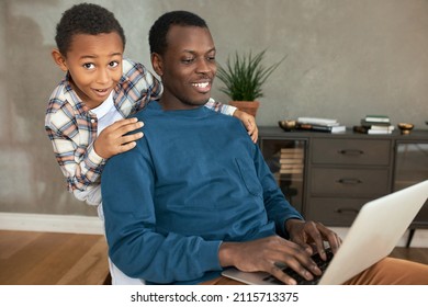 Father And Son. Surprised And Astonished Little African Boy With Short Curly Hair In Plaid Shirt Standing Behind Dad's Back, Holding Hands Over His Shoulder, While His Parent Working On Laptop