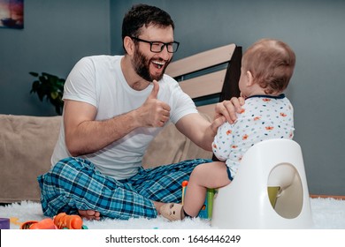 Father And Son. The Father Teaches The Child To Walk On The Potty. Father Is Very Happy And Shows Approx. Dad Put His Hand On The Baby Shoulder And Supports Him. Baby Toiletries. Potty Training