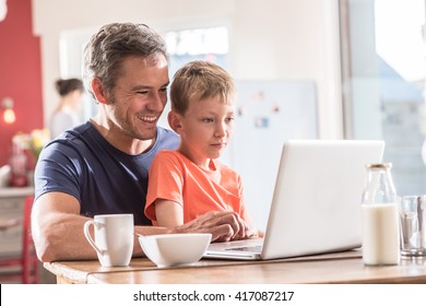 A father and son using a laptop while having breakfast, they are wearing casual clothes, the mother works in the blurred background