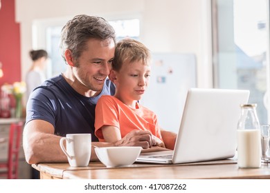 A father and son using a laptop while having breakfast, they are wearing casual clothes, the mother works in the blurred background