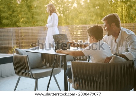 Father And Son Using Tablet On Home Balcony Terrace With Mother In Background Family Bonding