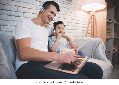 Father And Son Are Watching Movie On Laptop With Popcorn On Couch At Night At Home.