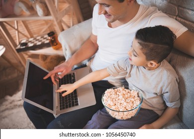 Father And Son Are Watching Movie On Laptop With Popcorn On Couch At Night At Home.