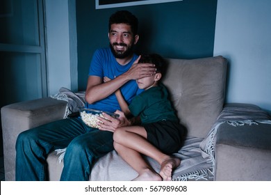 Father And Son Watching Scary Movie On Tv At Night. Father Covering His Son's Eyes