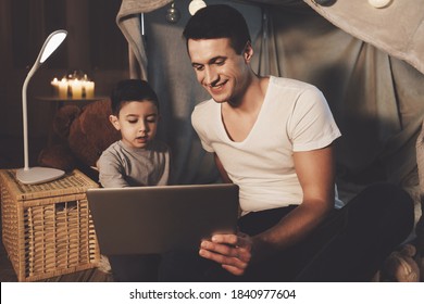 Father And Son Are Watching Video On Laptop In Blanket Fort At Night At Home. Dad And Little Son Are Sitting In A House And Watching A Movie On A Notebook.