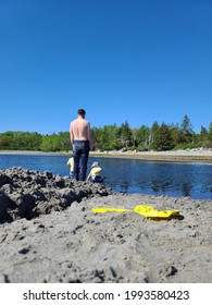 A Father And Sons Enjoying A Leisurely Afternoon At The Beach. They're Digging A Messy Moat And Their Plastic Shovel Is Laying Next To It. The Toddler Boys Are In The Water With Matching Swimsuits.