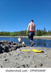 A Father And Sons Enjoying A Leisurely Afternoon At The Beach. They're Digging A Messy Moat And Their Plastic Shovel Is Laying Next To It. The Toddler Boys Are In The Water With Matching Swimsuits.