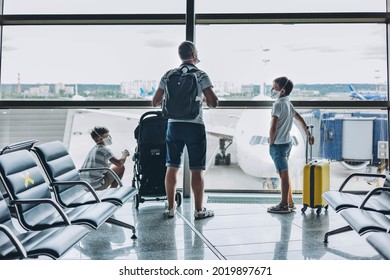 Father and sons in protective medical face mask with yellow suitcase at empty airport. Children looking out window at airplane. Family waiting for departure gate. Family trip and vacation concept.