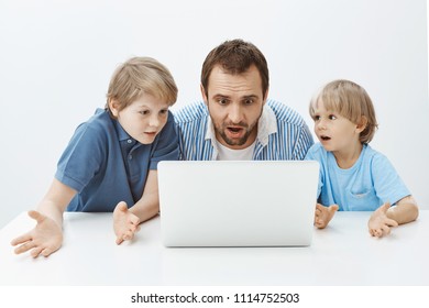 Father And Sons Watching Video Tutorial, Wanting Make Surprise For Mother. Portrait Of Clueless Confused Dad And Boys Sitting At Table And Looking At Laptop Screen, Being Unaware And Questioned