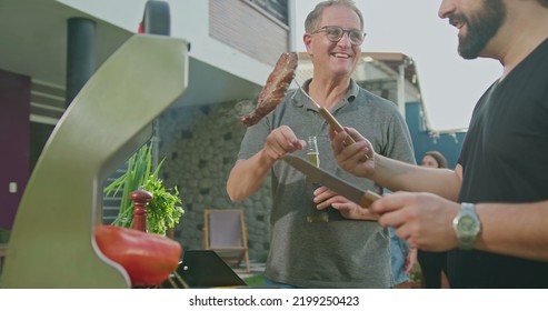 Father Standing By Son Grilling Meat. Adult Son Showing Steak To Dad. Friends And Family Gathered For Barbecue. Parent Talking To Son During Summer Holiday Celebration