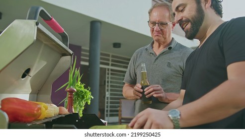 Father Standing By Son Grilling Meat. Adult Son Showing Steak To Dad. Friends And Family Gathered For Barbecue. Parent Talking To Son During Summer Holiday Celebration