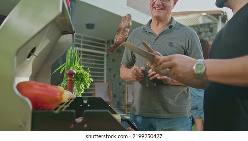 Father Standing By Son Grilling Meat. Adult Son Showing Steak To Dad. Friends And Family Gathered For Barbecue. Parent Talking To Son During Summer Holiday Celebration