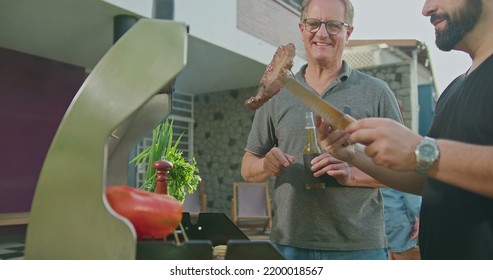 Father Standing By Son Grilling Meat. Adult Son Showing Steak To Dad. Friends And Family Gathered For Barbecue. Parent Talking To Son During Summer Holiday Celebration