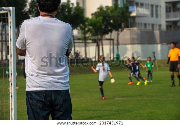 Father standing and watching his daughter\
playing football in a school tournament on a sideline with a sunny\
day. Sport, outdoor active, lifestyle, happy family and soccer mom\
and soccer dad concepts.