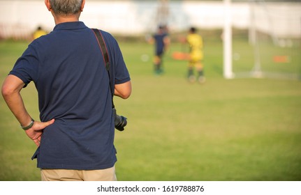 Father Standing And Watching His Son Playing Football In A School Tournament On A Clear Sky And Sunny Day. Sport, Outdoor Active, Lifestyle, Happy Family And Soccer Mom And Soccer Dad Concept.