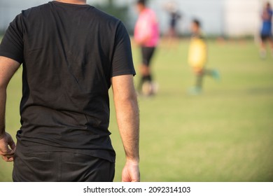 Father Standing And Watching His Son Playing Football In A School Tournament On A Sideline With A Sunny Day. Sport, Outdoor Active, Lifestyle, Happy Family And Soccer Mom And Soccer Dad Concept.