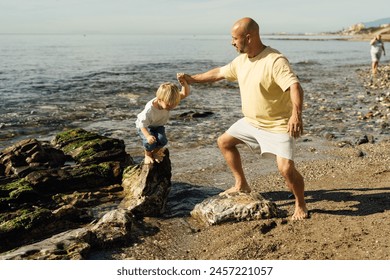 father supports son on rocks in the sea. Shot of father helping his son to jump over the rock on the beach. Young man and little boy having fun at the rocky beach. - Powered by Shutterstock