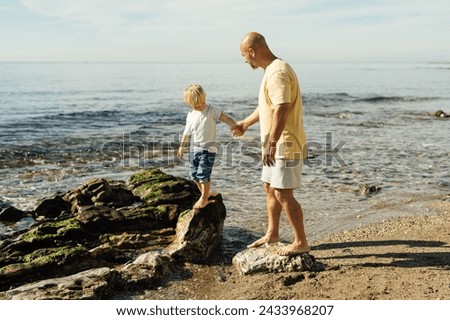 father supports son on rocks in the sea. Shot of father helping his son to jump over the rock on the beach. Young man and little boy having fun at the rocky beach.