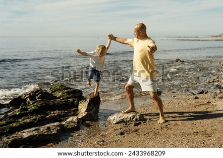 father supports son on rocks in the sea. Shot of father helping his son to jump over the rock on the beach. Young man and little boy having fun at the rocky beach.