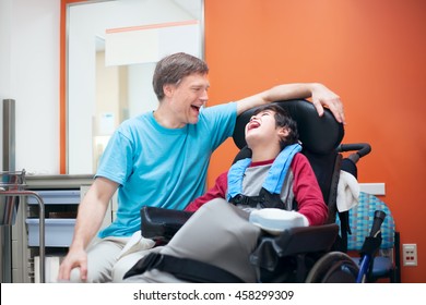 Father talking with disabled biracial son sitting in wheelchair while waiting in doctor's office, laughing together.