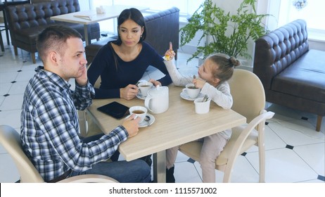 Father Talking To Phone While Sitting With His Wife And Daughter In Cafe Or Restaurant. Mother Looking Angry At Him.