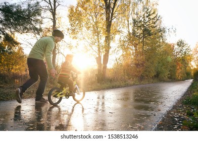 Father Teaches His Little Child To Ride Bike In Autumn Park. Happy Family Moments. Time Together Dad And Son. Candid Lifestyle Image. 