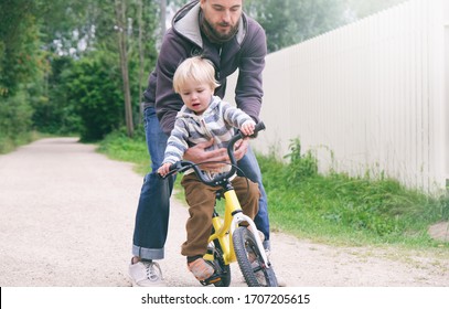 Father Teaches His Little Child To Ride Bike In Spring Summer Park, Catches Boy When He Falls. Happy Family Moments. Time Together Dad And Son. Candid Lifestyle Image.