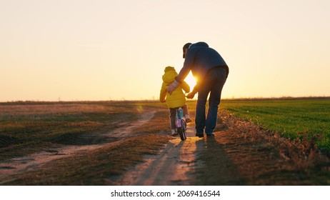 Father Teaches A Little Kid Girl To Ride A Child's Bike On The Road, In The Fall, In Spring. Happy Family, Childhood. Happy Family, Dad Teaches His Daughter, Child To Ride A Bike In The Park At Sunset