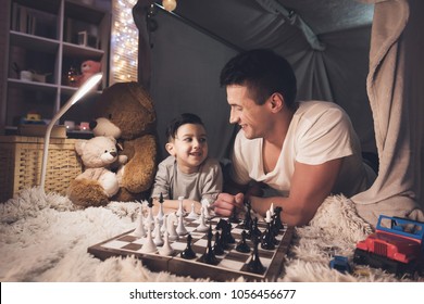 Father teaches little son how to play chess in blanket fort at night at home.