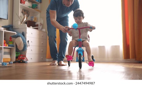 father teaches son baby to ride bike. happy family childhood dream concept. father and little son baby learn to ride a bike indoors home. happy family goes in for sunlight sports indoors - Powered by Shutterstock