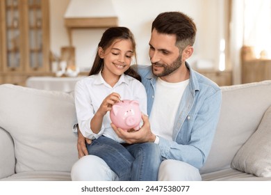 Father teaching daughter about savings, both smiling as they insert money into a piggy bank while seated - Powered by Shutterstock