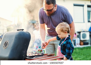 Father teaching his little son how to grill while standing in backyard at summer. Family gathering concept.