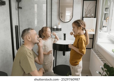 A father is teaching his two daughters how to brush their teeth in the bathroom. They are all smiling and enjoying the experience. The bathroom is clean and modern with white walls and black accents. - Powered by Shutterstock