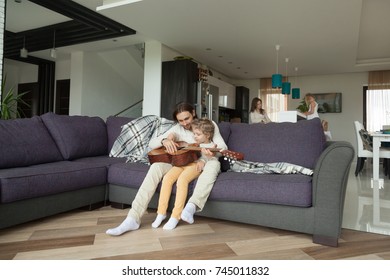 Father teaching little son to play guitar sitting on sofa while daughter helping mother on kitchen, young parents spending time with children at home, happy family and kids leisure activities - Shutterstock ID 745011832