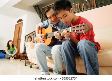 Father Teaching Son To Play Guitar, Mother And Daughter In The Background