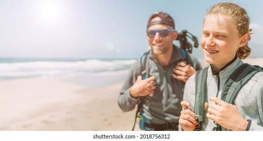 Father With Teenager Son With Backpacks Walking By The Sandy Seaside Beach. They Smiling And Looking At The Camera. Active Happy Family People Vacation Time Concept Image.