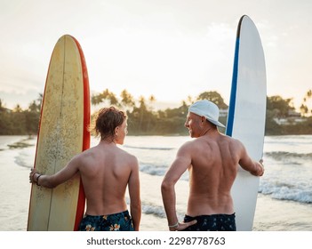 Father with teenager son standing with surfboards on the sandy ocean beach with palm trees on background lightened with sunset sun. They smiling and have a conversation. Family active vacation concept - Powered by Shutterstock