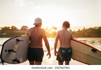 Father with teenager son walking with surfboards by the sandy ocean beach with palm trees on background lightened with sunset sun. They smiling and have a conversation. Family active vacation concept. - Powered by Shutterstock