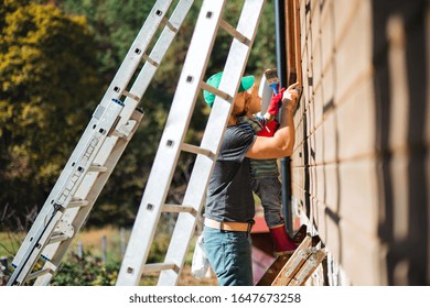 A Father And Toddler Boy Outdoors In Summer, Painting Wooden House.