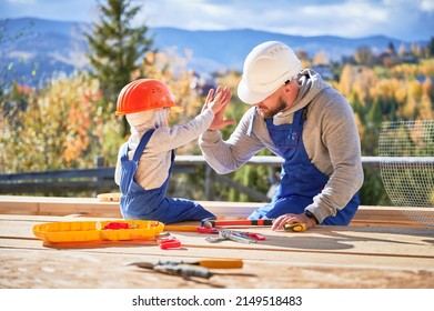 Father with toddler son building wooden frame house. Male builder giving high five to kid on construction site, wearing helmet and blue overalls on sunny day. Carpentry and family concept.