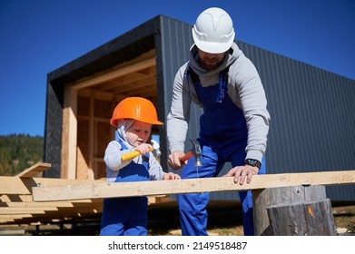 Father with toddler son building wooden frame house. Male builders hammering nail into plank on construction site, wearing helmet and blue overalls on sunny day. Carpentry and family concept.