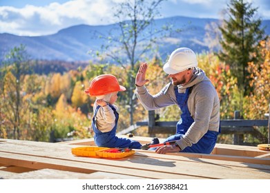 Father With Toddler Son Building Wooden Frame House. Male Builder Giving High Five To Kid On Construction Site, Wearing Helmet And Blue Overalls On Sunny Day. Carpentry And Family Concept.