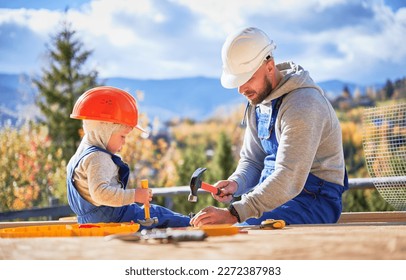 Father with toddler son building wooden frame house. Male builders hammering nail into plank on construction site, wearing helmet and blue overalls on sunny day. Carpentry and family concept. - Shutterstock ID 2272387983